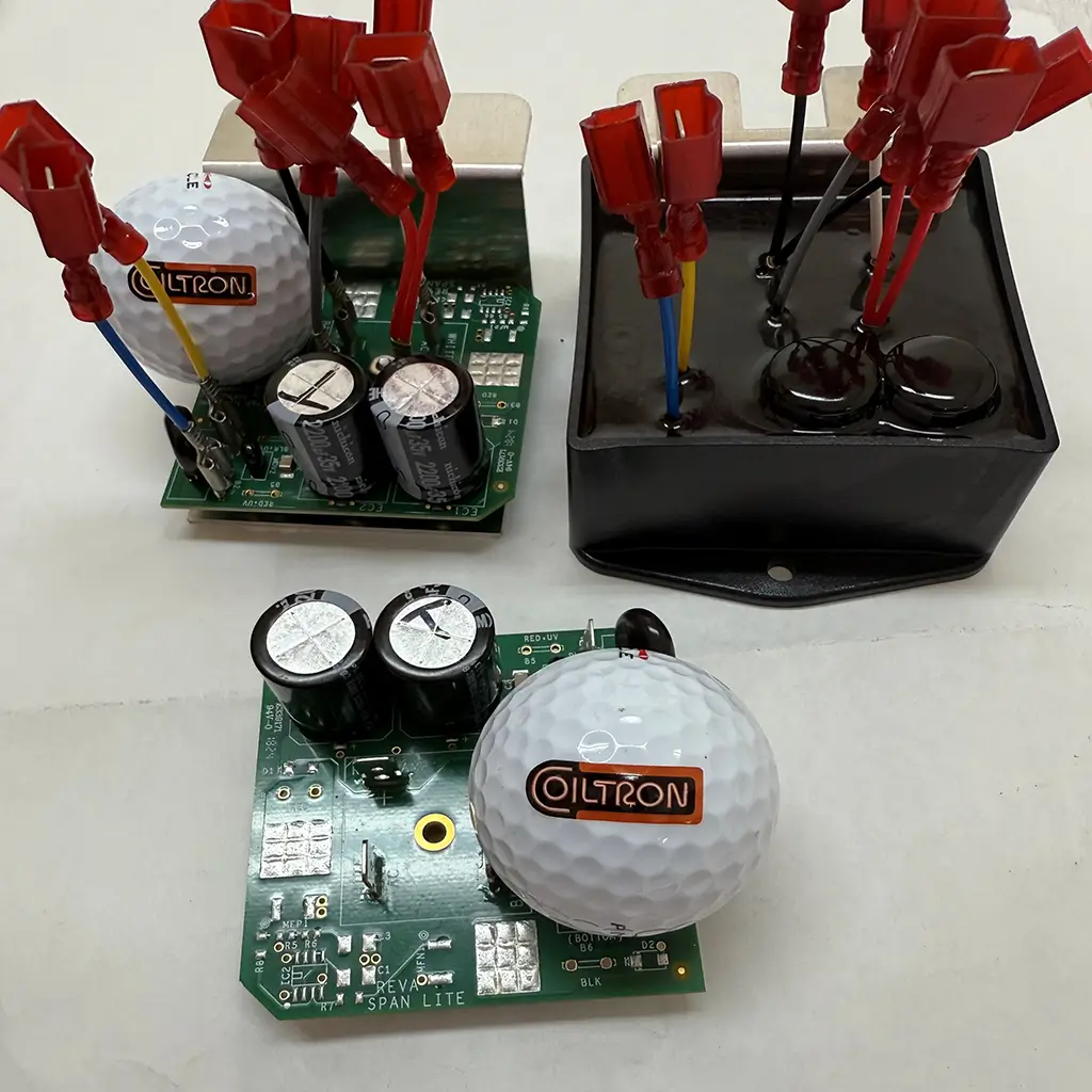 A trio of Coiltron custom electronics: two SPAN LITE Rev A PCBs with large capacitors and heat sinks, and one fully encapsulated black power module with multiple red quick-disconnect terminals, featuring golf balls for scale.