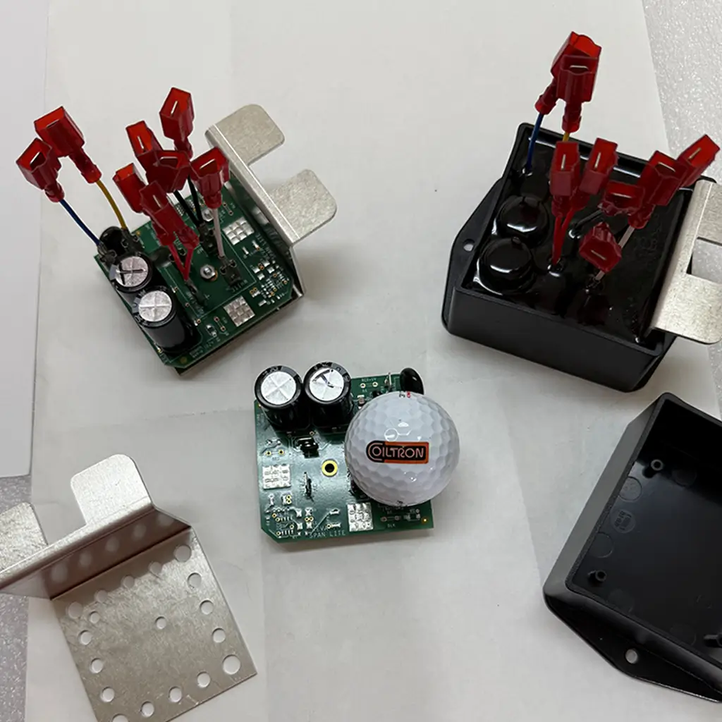 Coiltron custom PCB assemblies with electrolytic capacitors shown next to a separate metal heat sink, an empty black housing, and a final potted power module with red wire terminals.